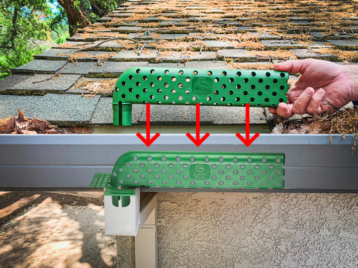 A person holding a green Gutter Stick above a house gutter filled with leaves and debris. Red arrows indicate the installation process, showing the Gutter Stick being placed into the downspout opening. The background features a shingled roof with pine needles and leaves. The installed Gutter Stick is visible in the gutter, demonstrating its placement and functionality in preventing clogs.