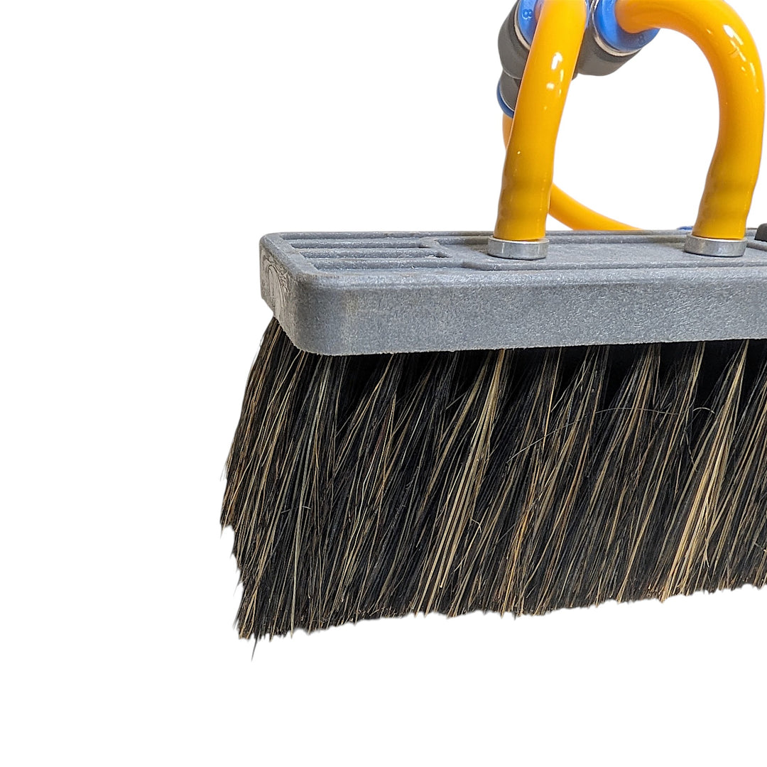 Close-up of a heavy-duty cleaning brush with black and brown bristles, a gray plastic handle plate, and bright yellow tubing on a white background.