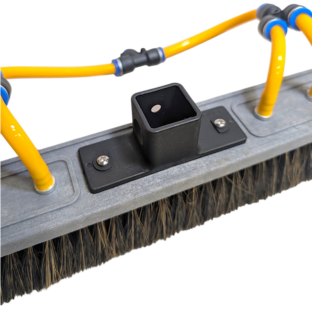Close-up of a heavy-duty cleaning brush attachment with a gray plastic frame, black mounting bracket, and bright yellow hoses connected via black and blue couplers, shown on a white background.