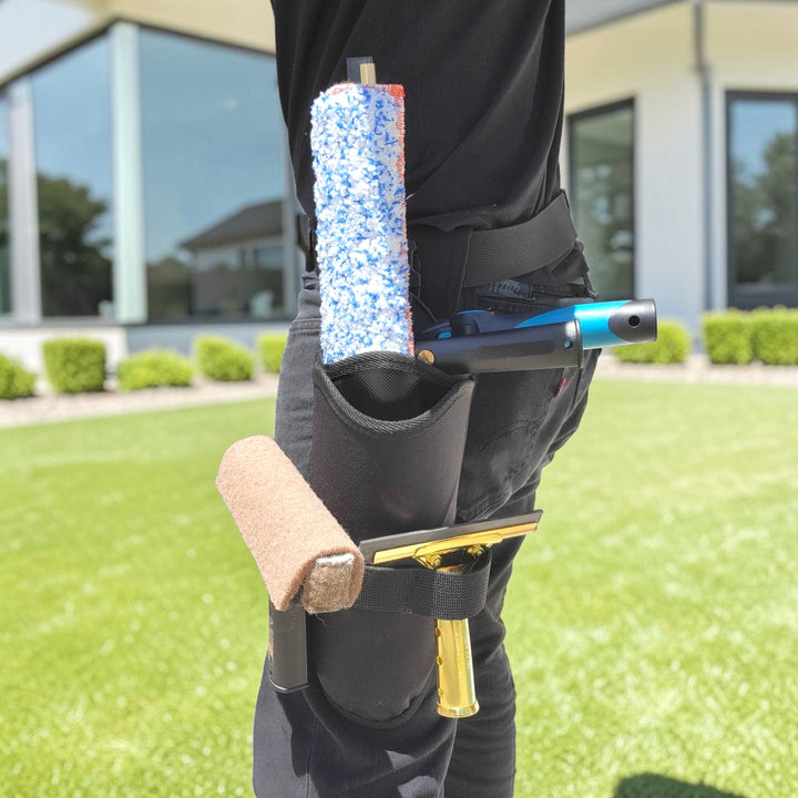 A person outdoors wearing a black utility belt with multiple cleaning tools attached, including a blue-and-white roller, a squeegee, and a brush, standing on a green lawn in front of a modern building.