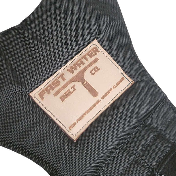 Close-up of a dark black textured fabric item with a rectangular tan patch reading 'FAST WATER BELT CO.' on a white background.