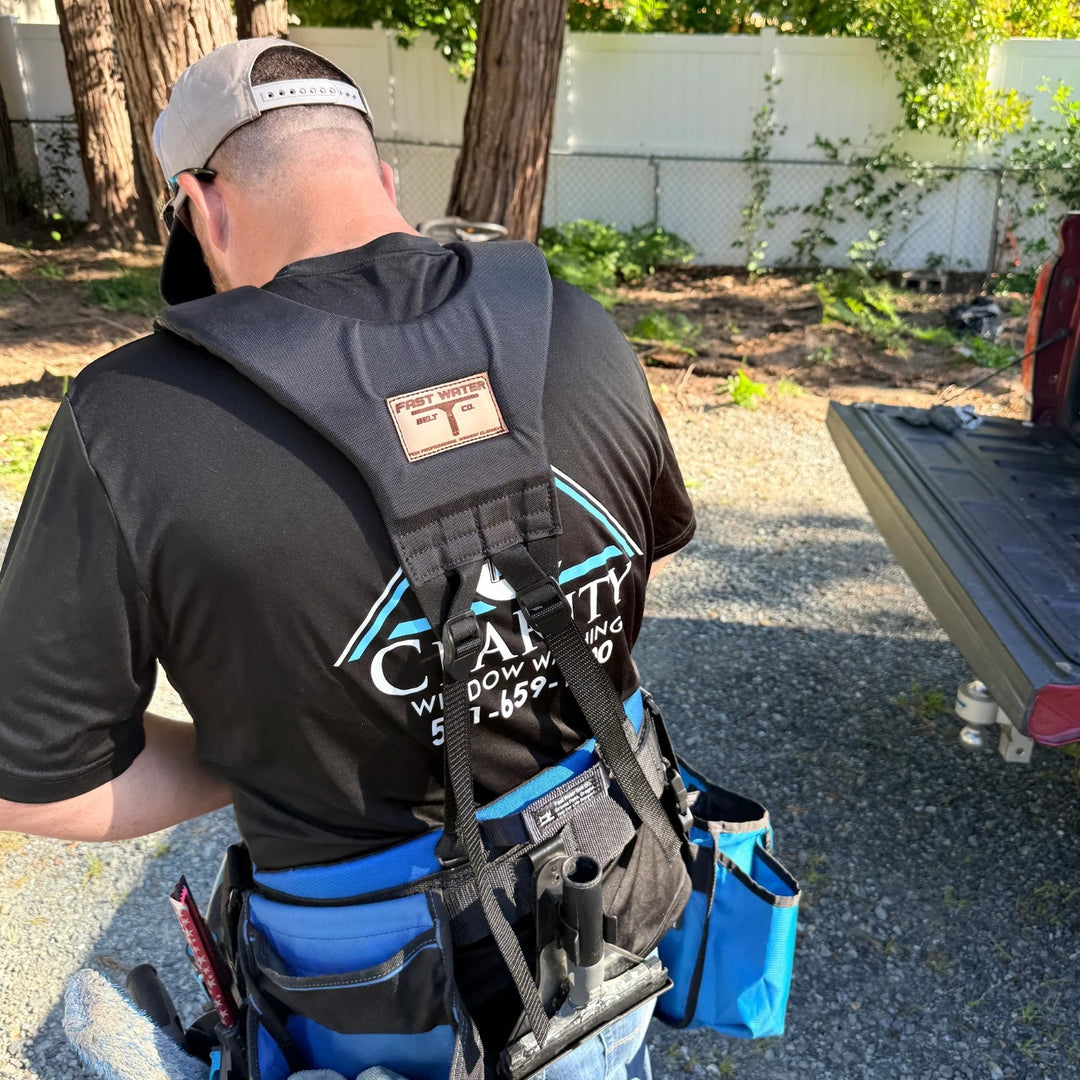A man viewed from behind wearing a black utility belt, and a black shoulder harness vest with a brand patch on the back, standing outdoors in a backyard setting.